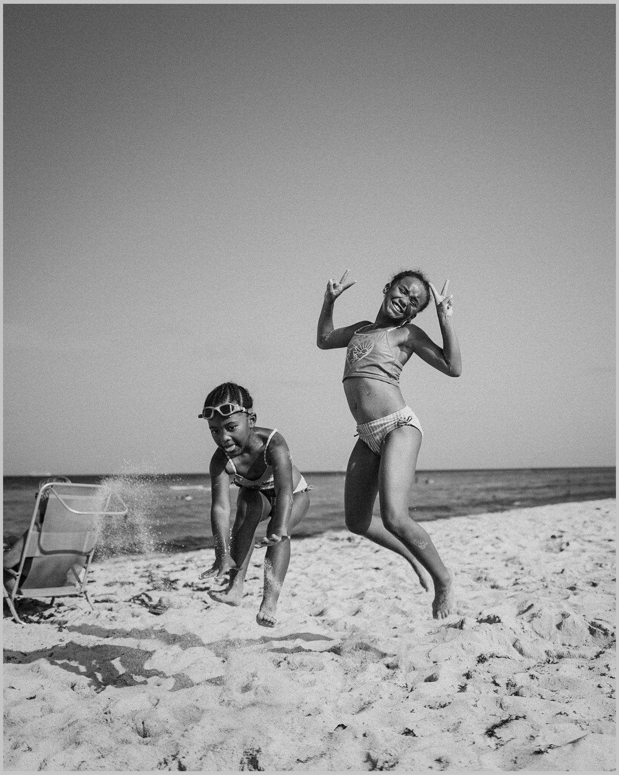Two girls playing on a sandy beach — one leaping into the air flashing peace signs, the other crouching to scoop up the sand